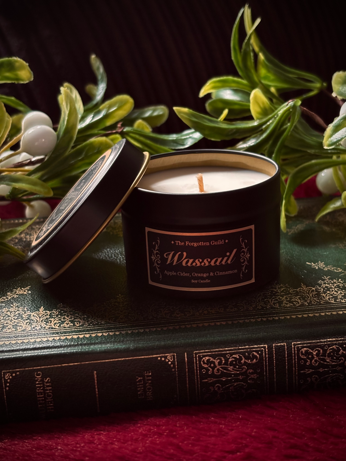 A black metal candle tin labeled 'Wassail' by The Forgotten Gold, shown open with cream-colored wax and a single wick, styled on top of an antique green book with decorative foliage and white berries around it.