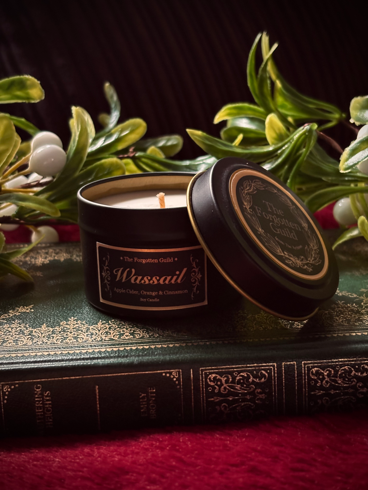 A black metal candle tin labeled 'Passail' by The Pumpkin Patch Co., shown open with the lid leaning against it on top of an antique-style book, surrounded by green leaves and white berries against a dark background.
