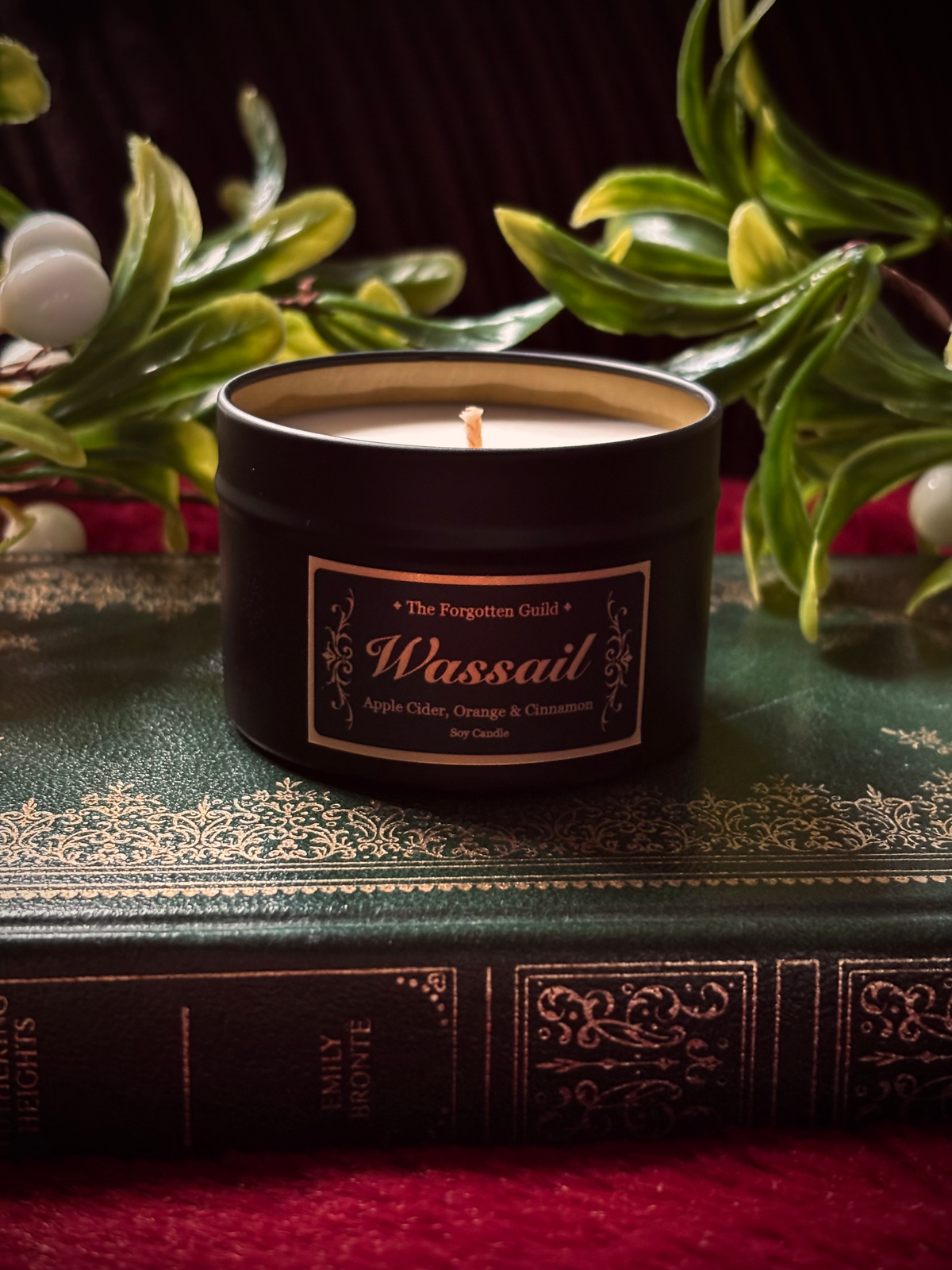 A black metal candle tin labeled 'Wassail' by The Forgotten Guild, styled on top of an antique green book with red velvet fabric and green foliage in the background.